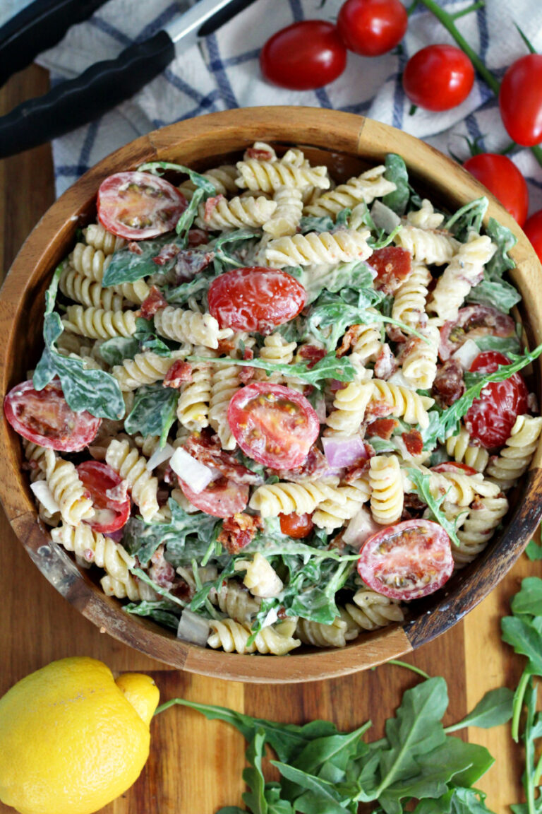Top view of BLT Pasta Salad in wooden serving bowl on top of wooden board. Lemon, arugula, cherry tomatoes, tongs, and kitchen towel also in partial view.