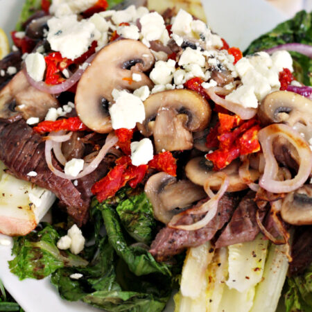 Close up of Steak and Blue Cheese Grilled Salad topped with sliced steak, grilled onions, sun-dried tomatoes, cooked mushrooms, and crumbled blue cheese. Lemons, tongs, and fresh rosemary in partial view.