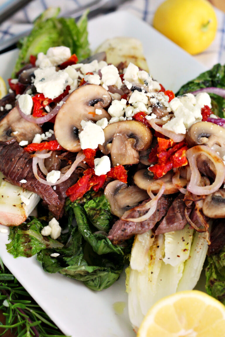 Close up of Steak and Blue Cheese Grilled Salad topped with sliced steak, grilled onions, sun-dried tomatoes, cooked mushrooms, and crumbled blue cheese. Lemons, tongs, and fresh rosemary in partial view.