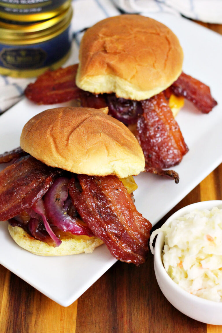 Memphis Burgers with Candied Bacon sit on white serving dish on top of wooden board. Small bowl of coleslaw, containers of seasoning, and kitchen towel also in partial view.