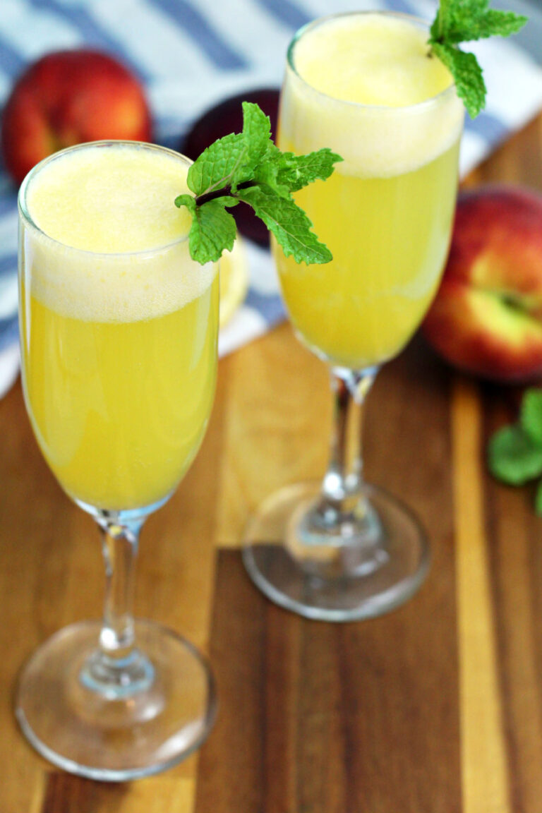 Two Peach Bellini mocktails sit on wooden board. Peaches and fresh mint sprigs in partial view in background.