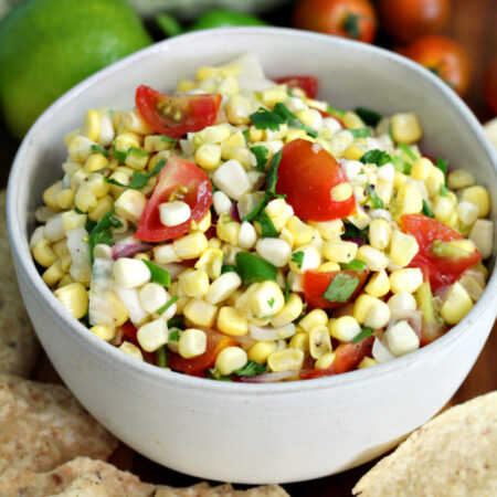 Corn and tomato salsa in small serving bowl sits on wooden board. Tortilla chips, limes, corn, and cherry tomatoes in partial view.