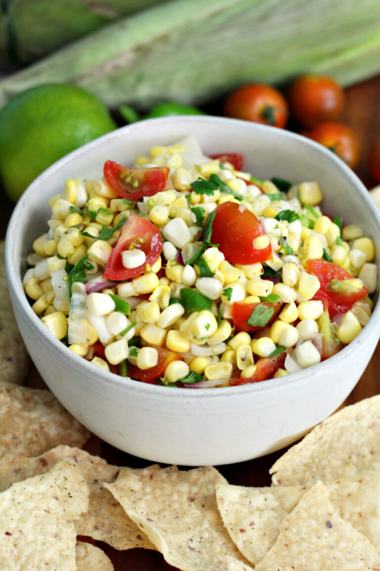 Corn and tomato salsa in small serving bowl sits on wooden board. Tortilla chips, limes, corn, and cherry tomatoes in partial view.