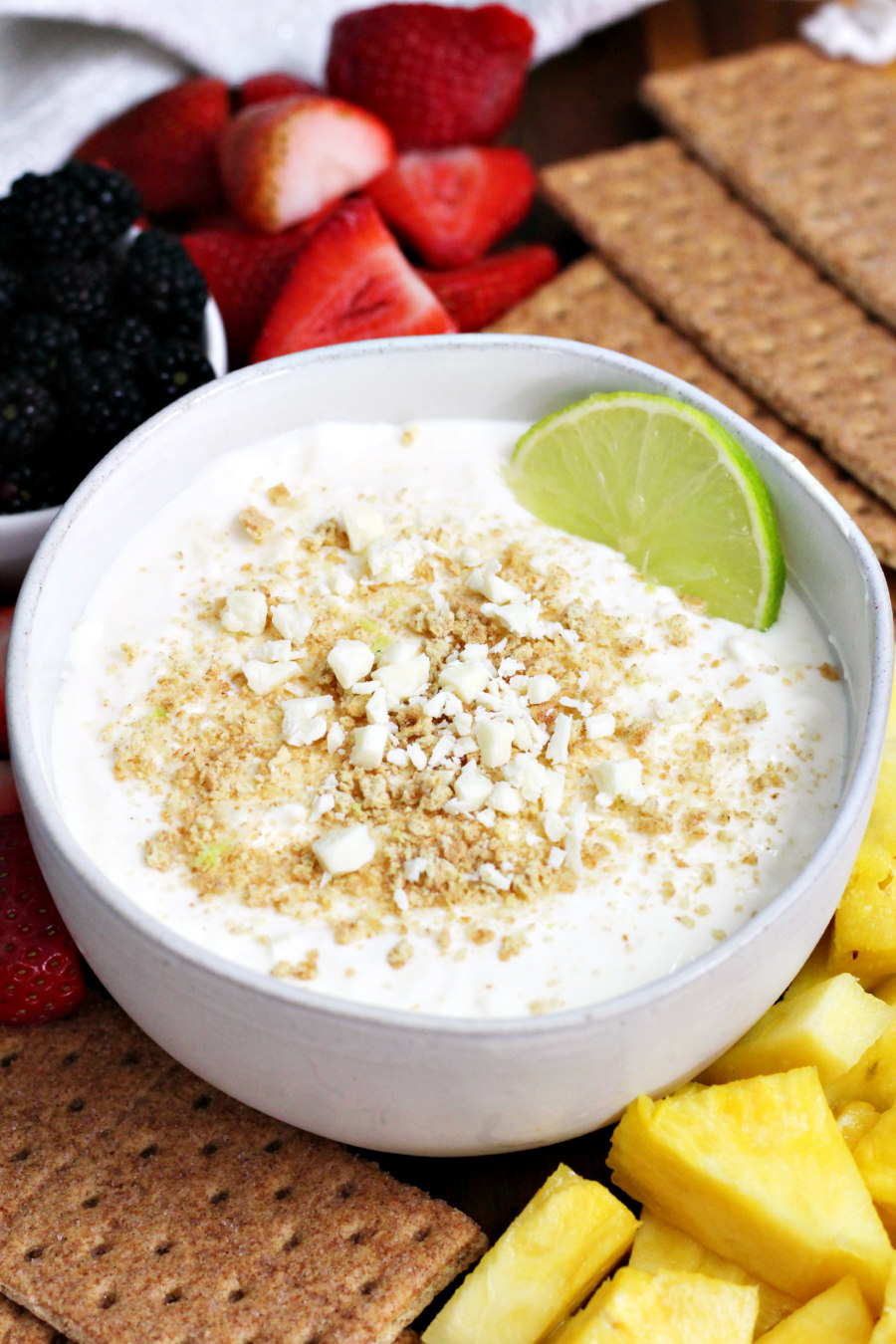 Healthier Key Lime Pie Dip served in serving bowl. Dish sits on wooden board surrounded by graham crackers and fresh blackberries, strawberries, and pineapple chunks.