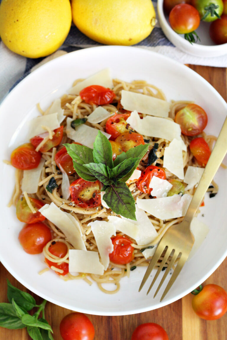 Top view of Tomato, Basil, and Lemon Spaghetti in serving dish with gold fork. Dish sits on top of wooden board. Spaghetti is garnished with shaved Parmesan cheese and fresh basil leaves. Lemons, cherry tomatoes, and fresh basil in partial view.