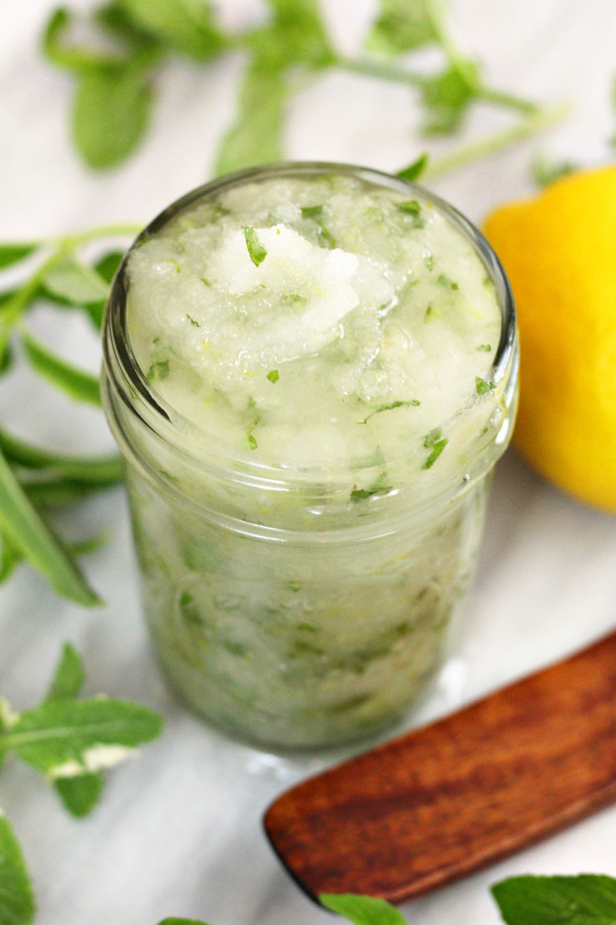 DIY Citrus-Mint Sugar Scrub in glass jar sits on marble board with fresh herb sprigs, lemon, and wooden spatula spoon.