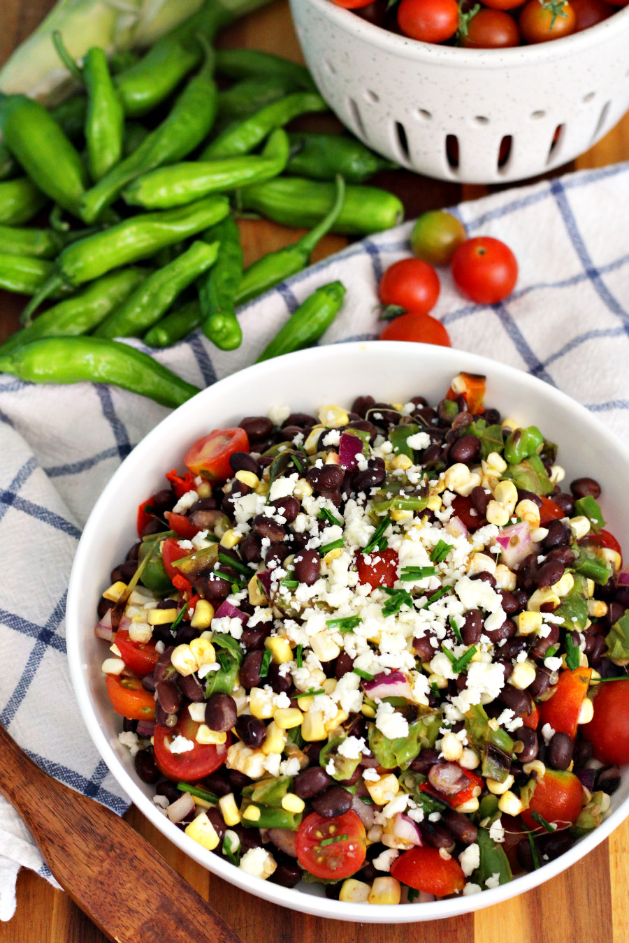 Shishito and Corn Salad with Black Beans sits in white serving dish. Dish sits on wooden board along with wooden spatula spoon, shishitos, cherry tomatoes, and ceramic colander filled with tomatoes.
