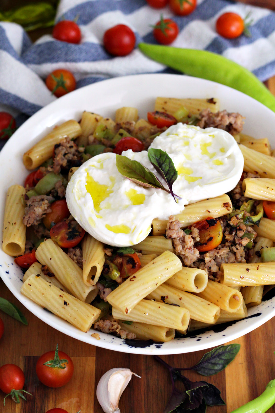 Sausage and Banana Pepper Rigatoni with Burrata sits in serving dish on wooden board. Cherry tomatoes, garlic clove, basil, and banana peppers also in partial view.