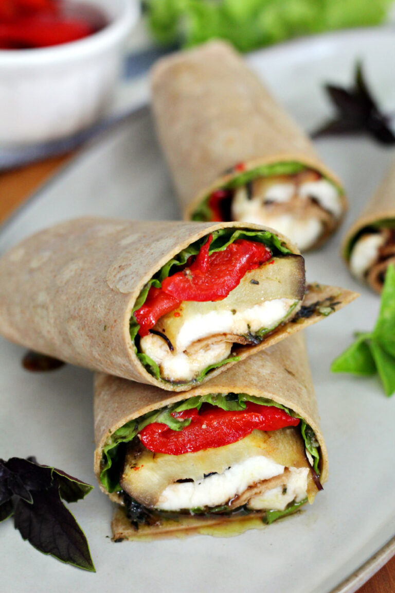Eggplant and Roasted Red Pepper Wrap sits on serving plate, cut in half, and on top of wooden board. Basil leaves are arranged on the plate and roasted red peppers and lettuce in partial view in background.
