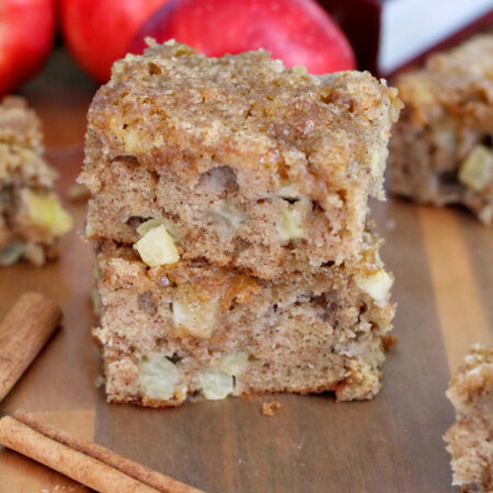 Two pieces of Apple Spice Snack Cake sit on wooden board. Cinnamon sticks, other pieces of cake, apples, and cookbook also in partial view.