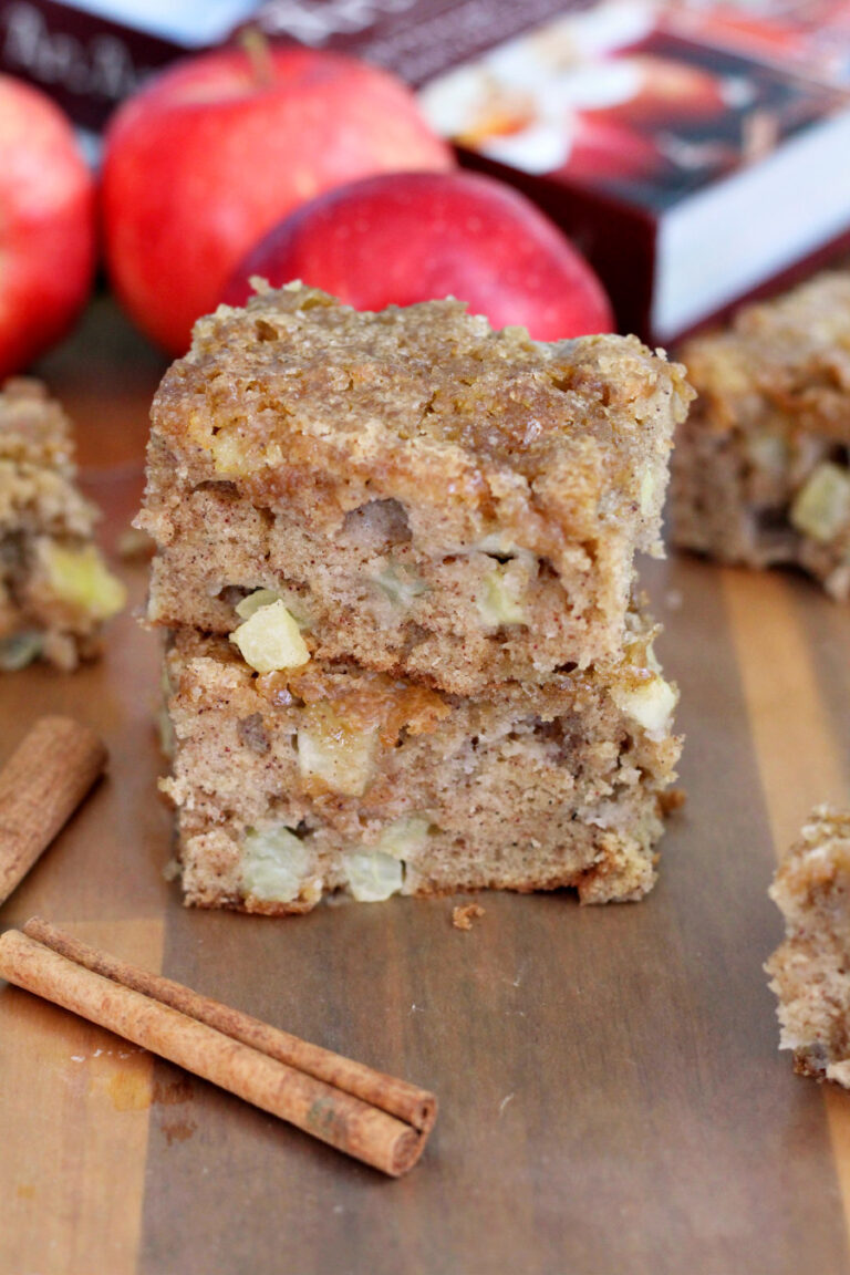 Two pieces of Apple Spice Snack Cake sit on wooden board. Cinnamon sticks, other pieces of cake, apples, and cookbook also in partial view.