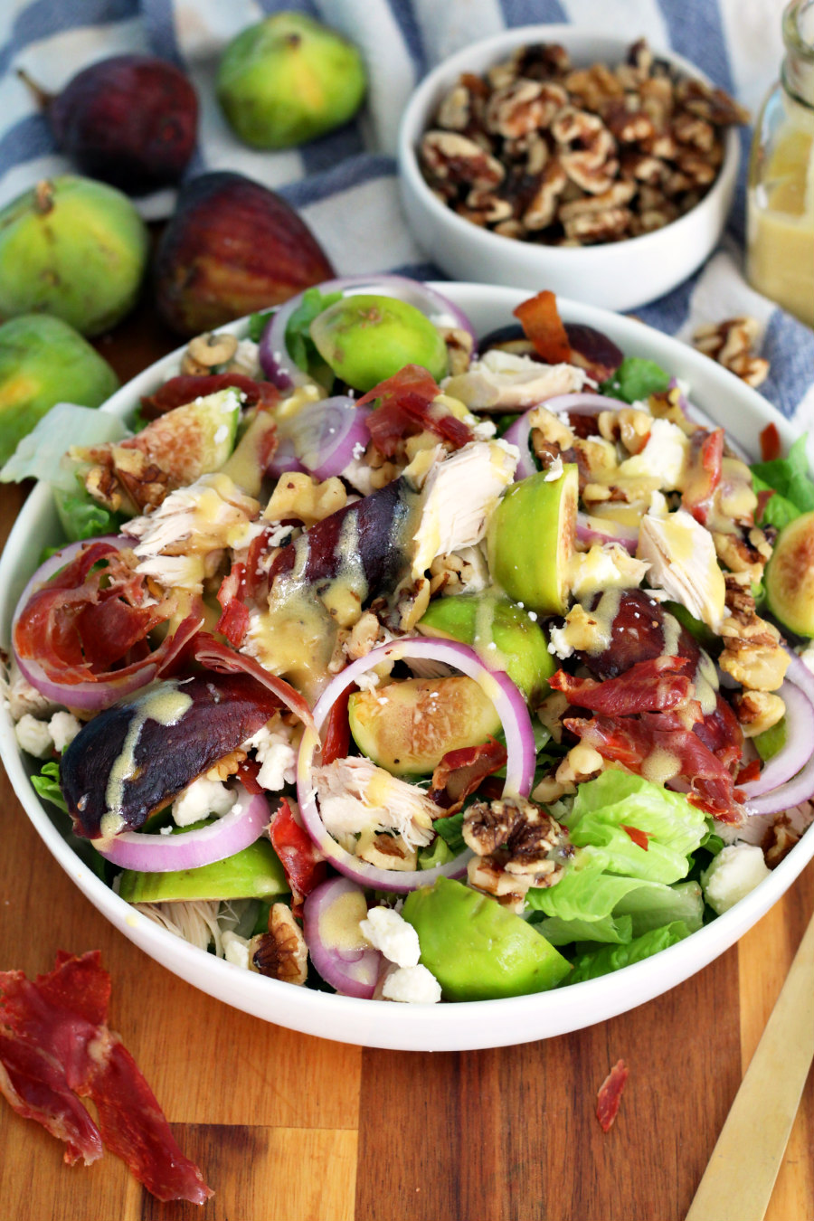 Harvest Chicken and Fig Salad sits in white serving dish on wooden board. Crispy prosciutto pieces, fresh figs, small bowl of walnuts, and gold fork in partial view.