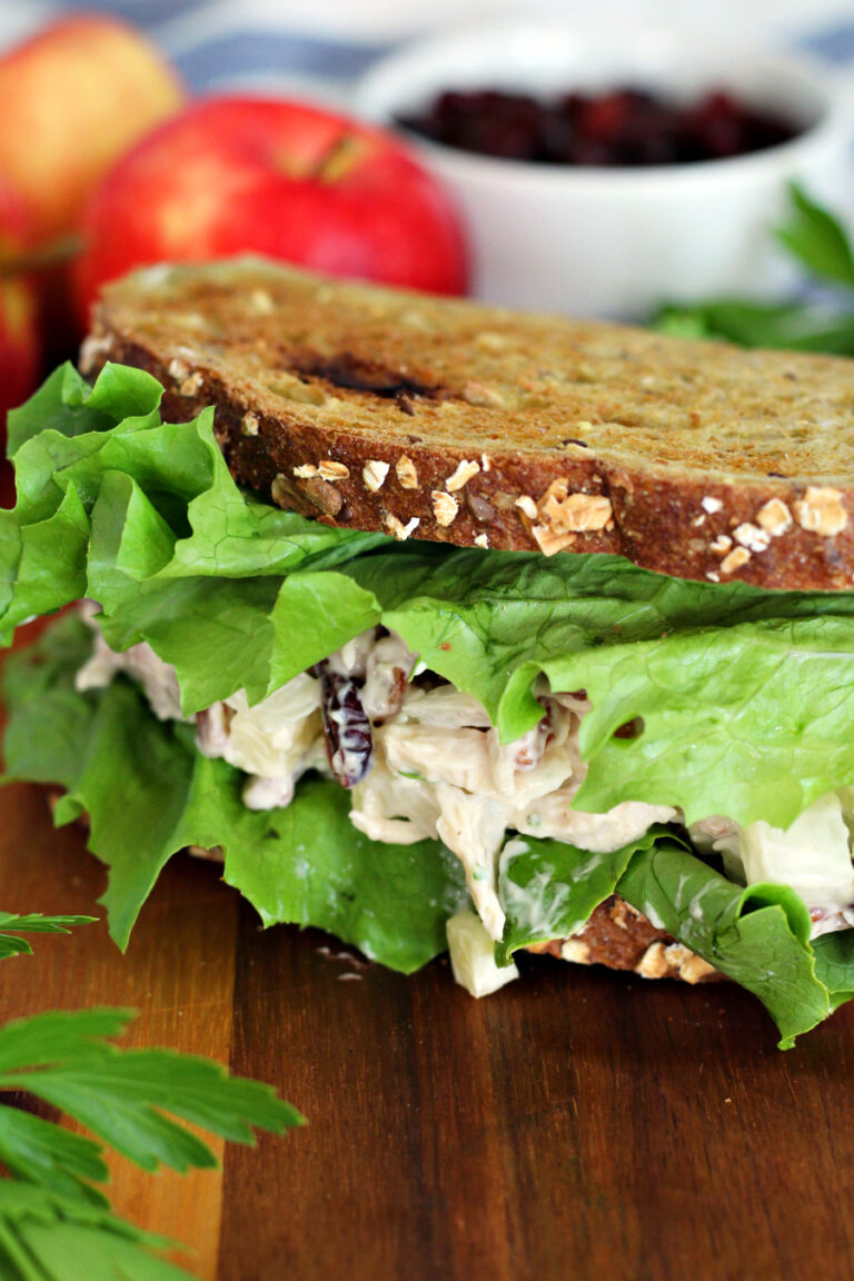 Partial view of Apple Pecan Chicken Salad sandwich on wooden board. Apples, fresh parsley, and small bowl of dried cranberries in partial view in background.