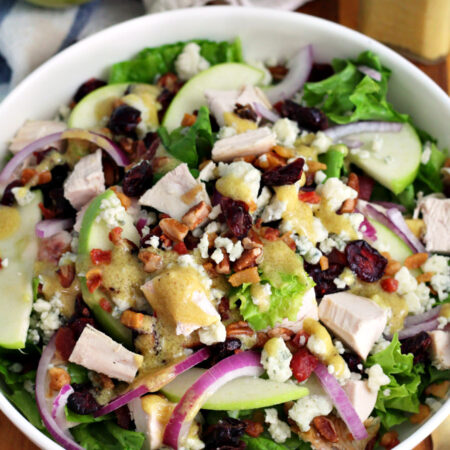 Chicken, Apple, and Blue Cheese Salad with Pancetta in white serving dish sits on wooden board. Dried cranberries, gold fork, Granny Smith apple, small bowl of chopped pecans, and small bottle of Dijon mustard dressing also in partial view.