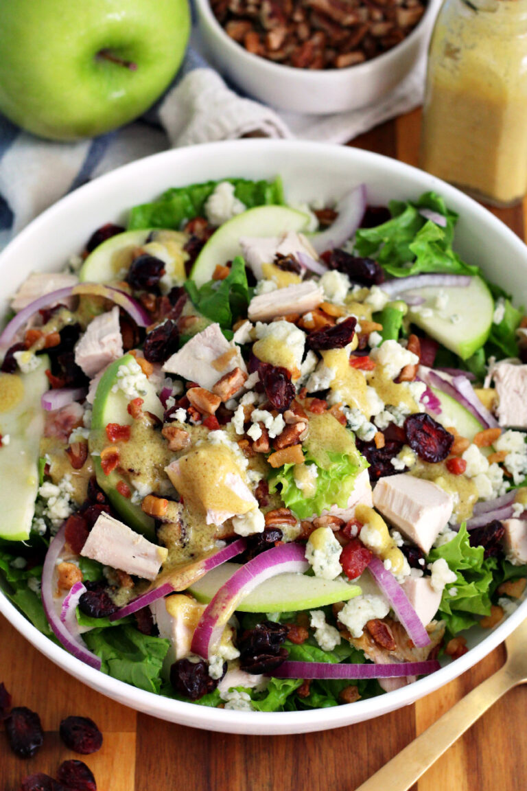 Chicken, Apple, and Blue Cheese Salad with Pancetta in white serving dish sits on wooden board. Dried cranberries, gold fork, Granny Smith apple, small bowl of chopped pecans, and small bottle of Dijon mustard dressing also in partial view.
