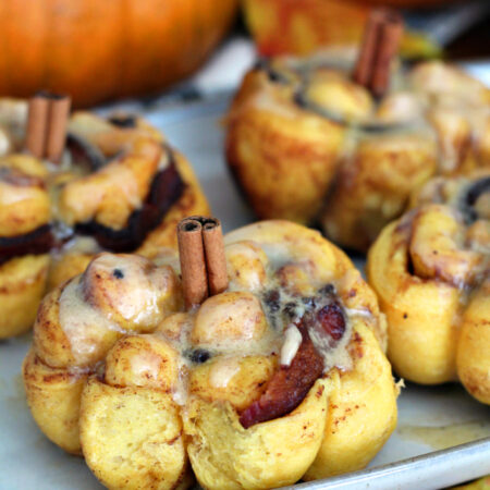 Pumpkin Spice Bacon Cinnamon Rolls sit on serving dish on wooden board. Pumpkins in partial view in background.