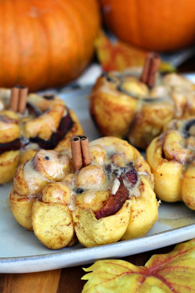 Pumpkin Spice Bacon Cinnamon Rolls sit on serving dish on wooden board. Pumpkins in partial view in background.