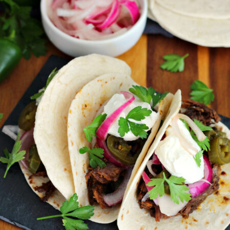 Angled view of Braised Short Rib Tacos on slate slab on top of wooden board. Tacos are in soft flour tortillas and dressed with pickled onions, pickled jalapenos, fresh parsley leaves, and sour cream. Fresh parsley, small bowl of pickled onions, and soft tortillas in partial view in background.