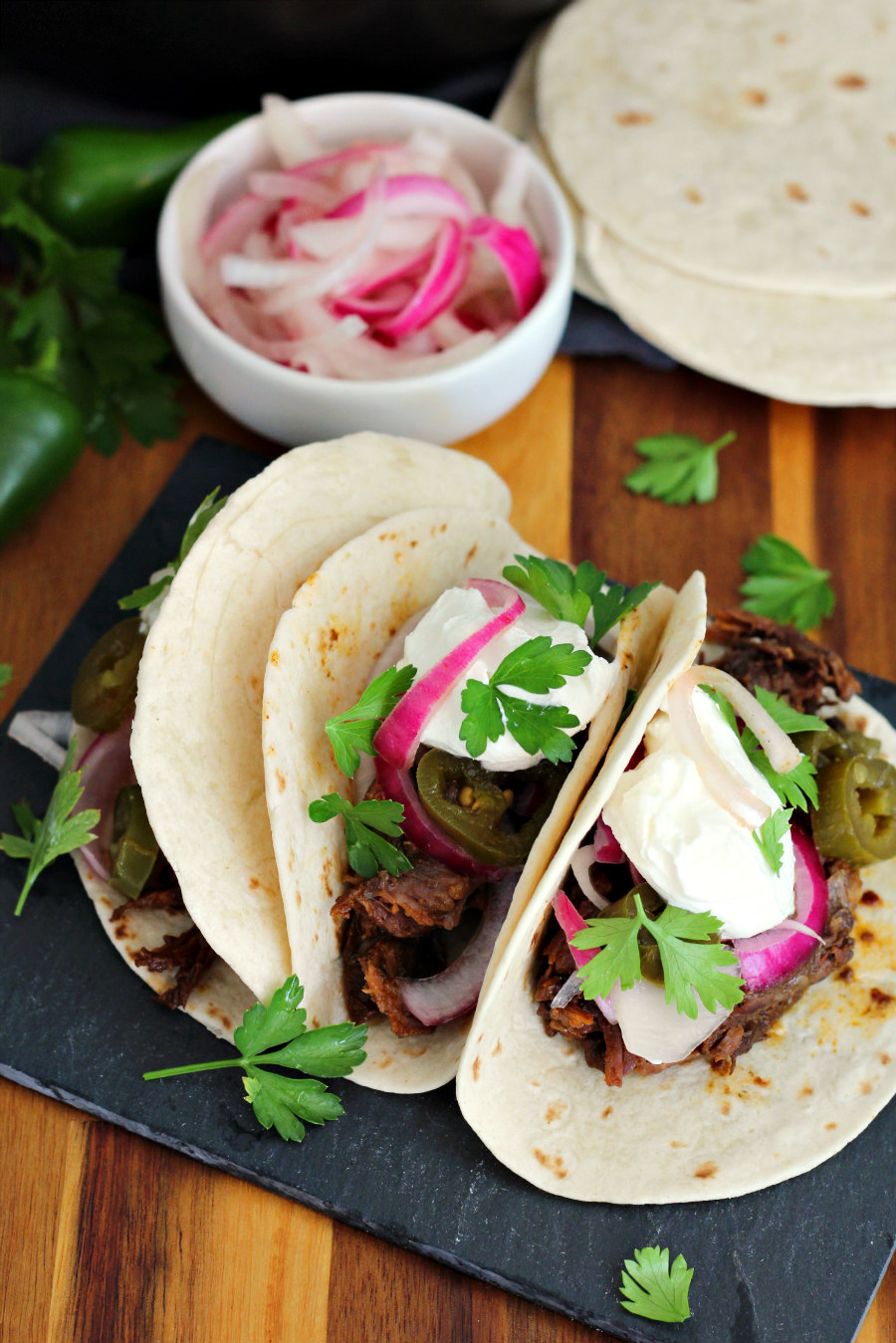 Angled view of Braised Short Rib Tacos on slate slab on top of wooden board. Tacos are in soft flour tortillas and dressed with pickled onions, pickled jalapenos, fresh parsley leaves, and sour cream. Fresh parsley, small bowl of pickled onions, and soft tortillas in partial view in background.
