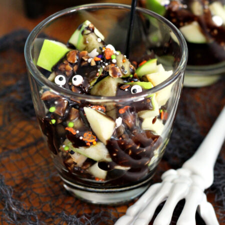 Deconstructed Halloween Caramel Apple Cup sits on wooden board with caramel apple cup in partial view in background. Decorative cauldron and skeleton hand also in partial view.