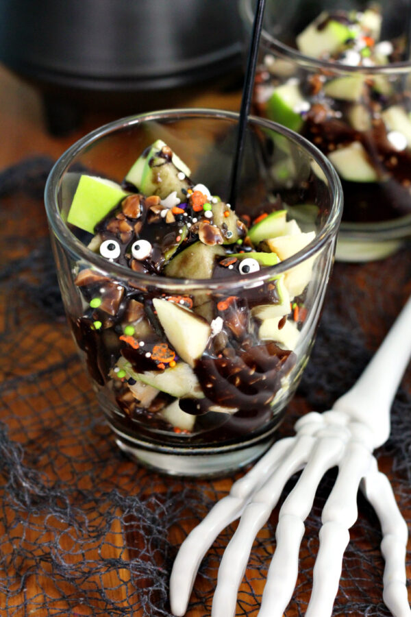 Deconstructed Halloween Caramel Apple Cup sits on wooden board with caramel apple cup in partial view in background. Decorative cauldron and skeleton hand also in partial view.