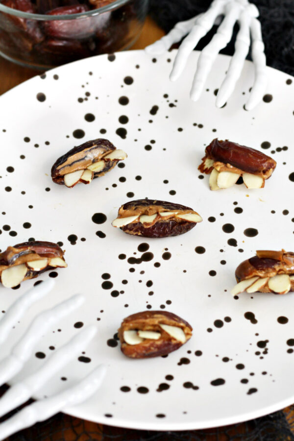 Almond Butter Stuffed Monster Dates sit on black speckled plate. Skeleton hands in partial view around plate. Small bowl of pitted dates in partial view in background.