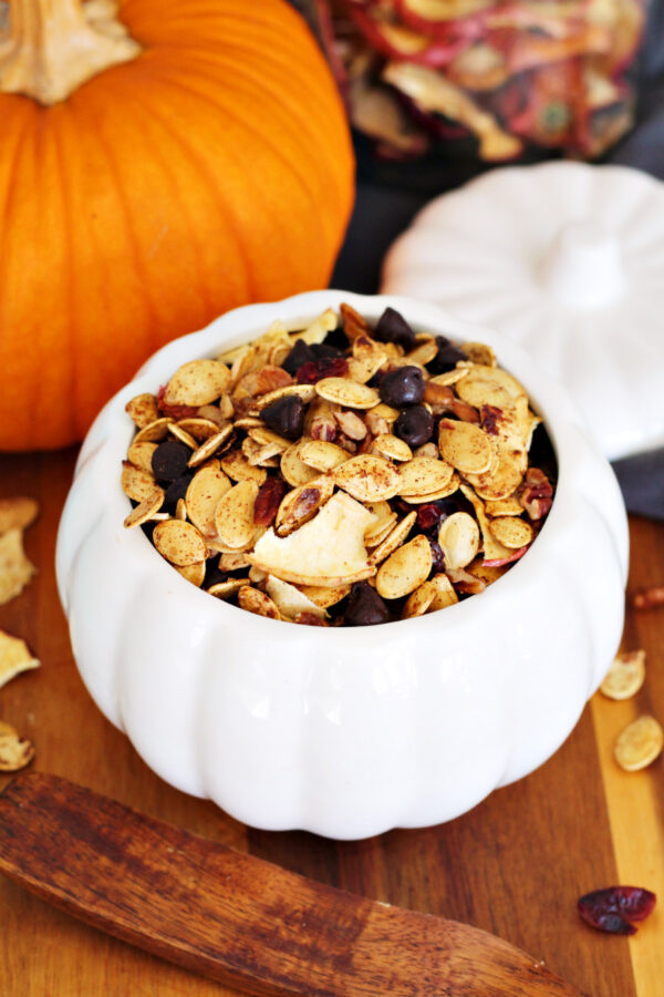 Roasted Pumpkin Seed Trail Mix sits inside white pumpkin shaped bowl on wooden board. Wooden spatula spoon, small pumpkin, and jar of dried apples in partial background.