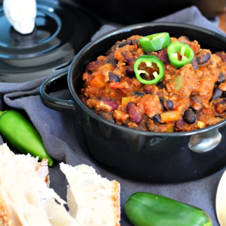 Samhain Pumpkin Chili with Ground Beef sits in black serving bowl on top of wooden board. Skeleton lid, cast iron pot of chili, jalepeños, and bread slices also in partial view.