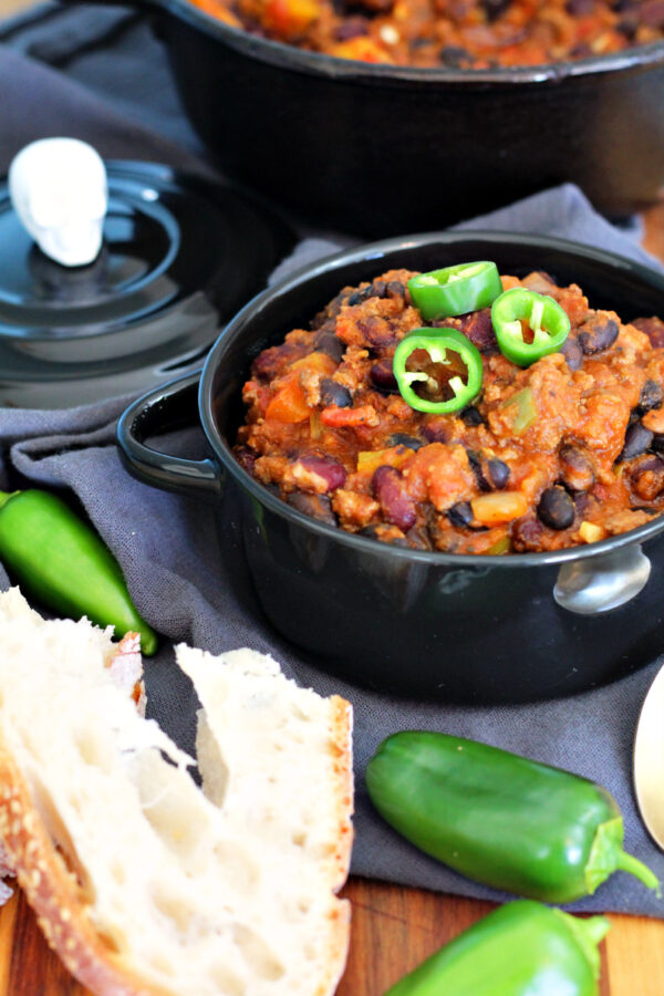 Samhain Pumpkin Chili with Ground Beef sits in black serving bowl on top of wooden board. Skeleton lid, cast iron pot of chili, jalepeños, and bread slices also in partial view.