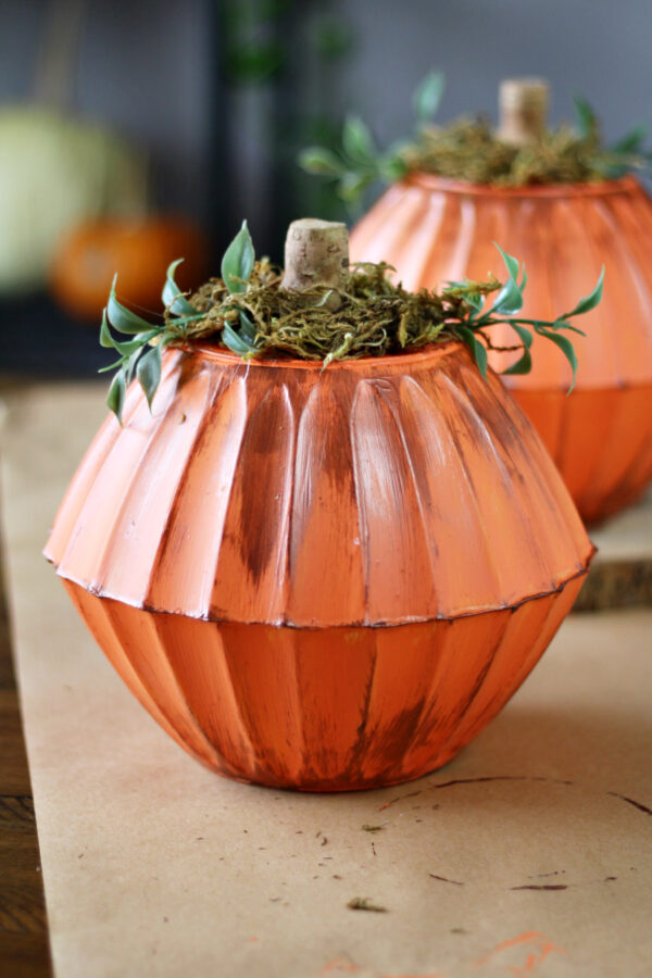 Dollar store bowl pumpkin sits on brown paper. Another dollar store bowl pumpkin sits in background on top of wood slice. Real pumpkins in partial blurred view in left side of background.