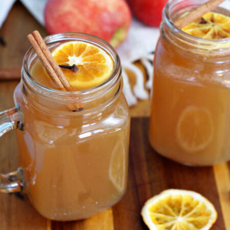 Glasses of Warm Maple Whiskey Apple Cider sit on top of wooden board. Orange slice, cloves, cinnamon sticks, and apples also in partial view.