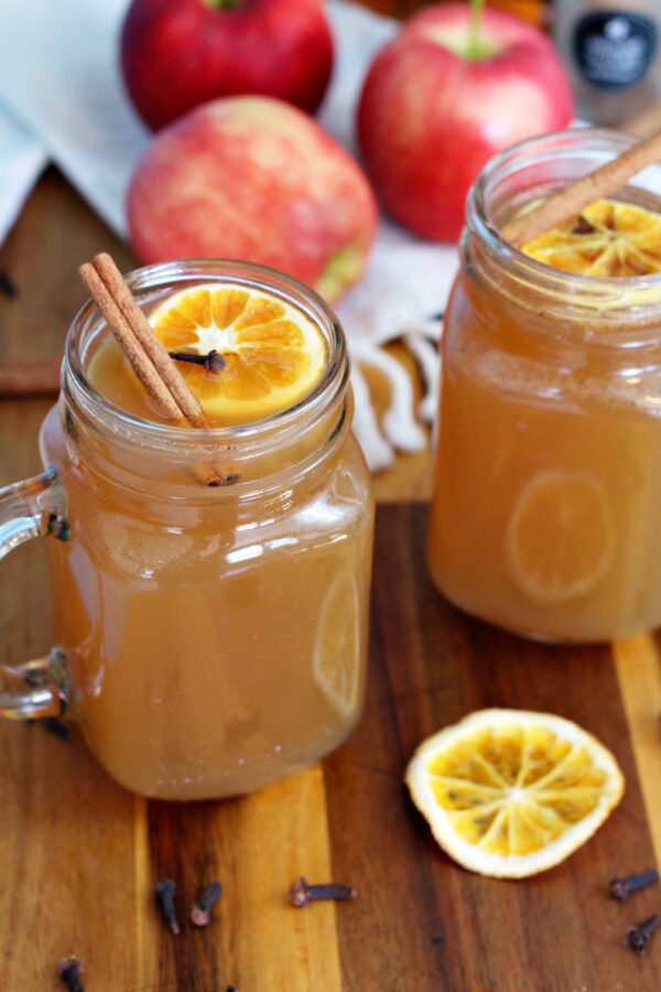 Glasses of Warm Maple Whiskey Apple Cider sit on top of wooden board. Orange slice, cloves, cinnamon sticks, and apples also in partial view.