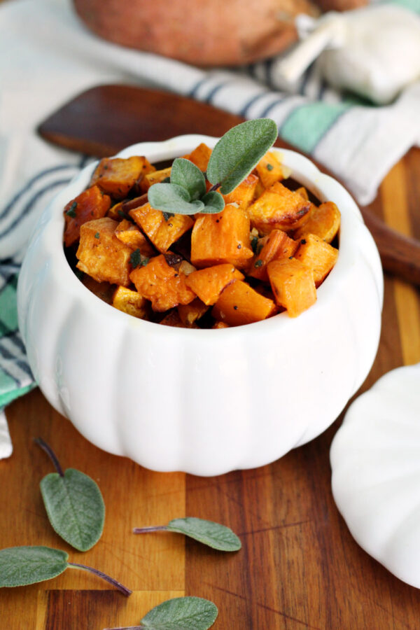 Roasted Sweet Potatoes with Sage Butter are served in pumpkin shaped dish. Dish sits on wooden board. Dish lid, fresh sage leaves,, wooden spatula spoon, garlic bulb, and sweet potato also in partial view.