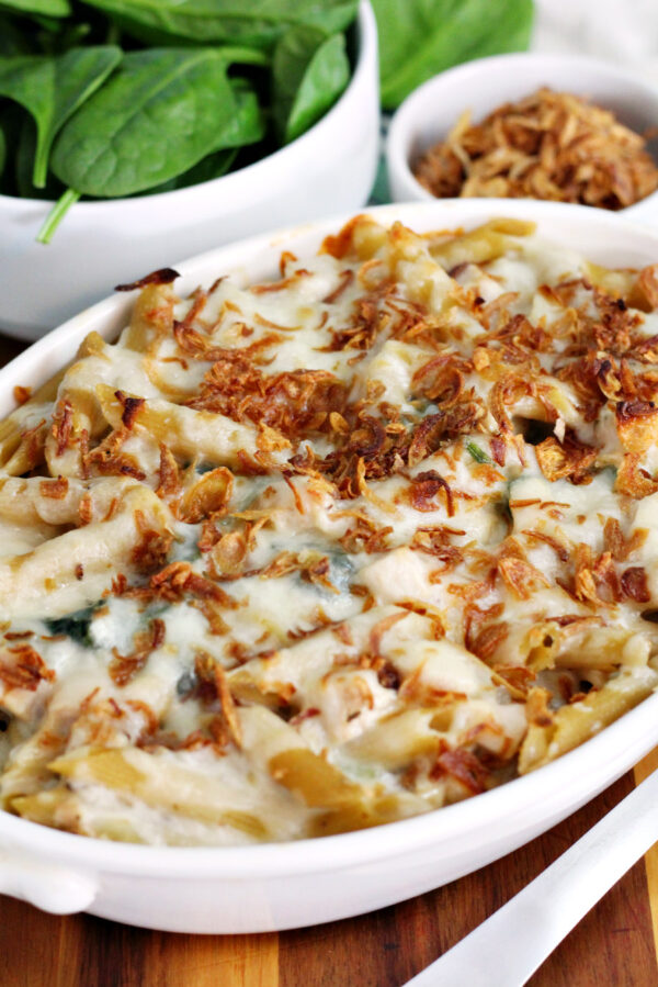 Leftover Turkey and Gravy Pasta Bake with Baby Spinach sits in serving dish on top of wooden board. Bowl of baby spinach, crispy fried onions, and metal spoon also in partial view.