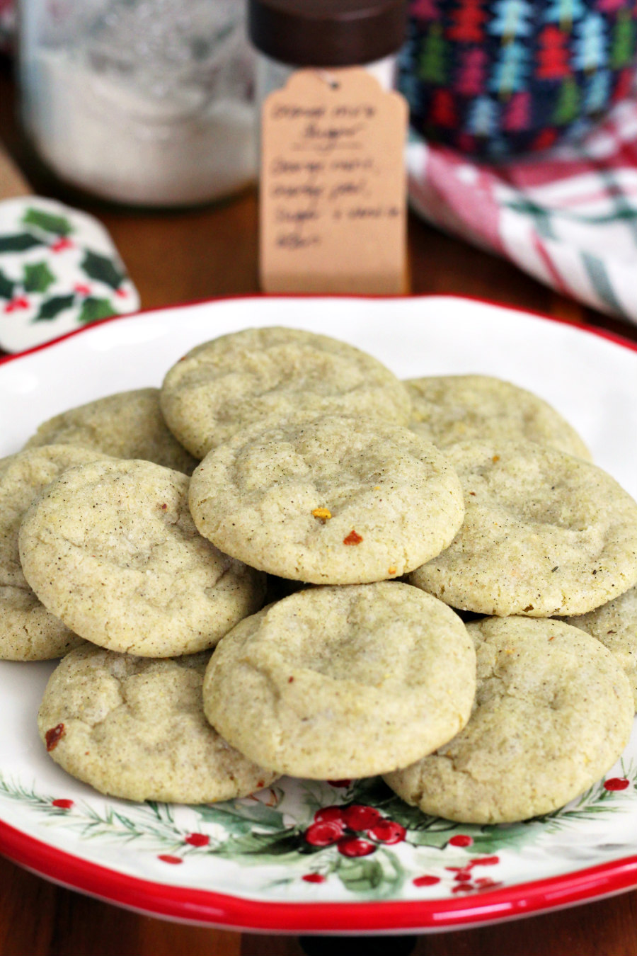 Orange Mint Vanilla Infused Sugar Cookies sit on holiday plate on top of wooden board. Holiday rubber spatula, jars of infused sugar, holiday tree bowl, and plaid Christmas towel in partial view in background.