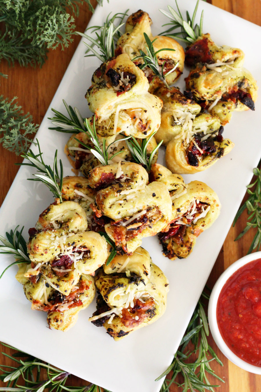 Top view of Pesto and Prosciutto Puff Pastry Christmas Trees on white serving dish. Dish sits on wooden board and is surrounded by greenery. A small bowl of marinara sauce is in partial view in the bottom right corner.