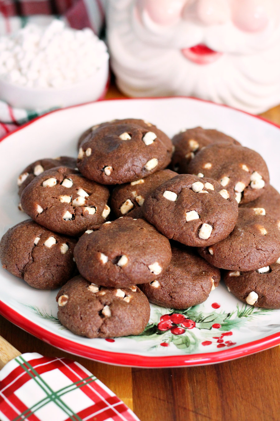 Easy Hot Cocoa Cookies sit on holiday plate on top of wooden board. Holiday rubber spatula, small bowl of mini marshmallows, and Santa container also in partial view.