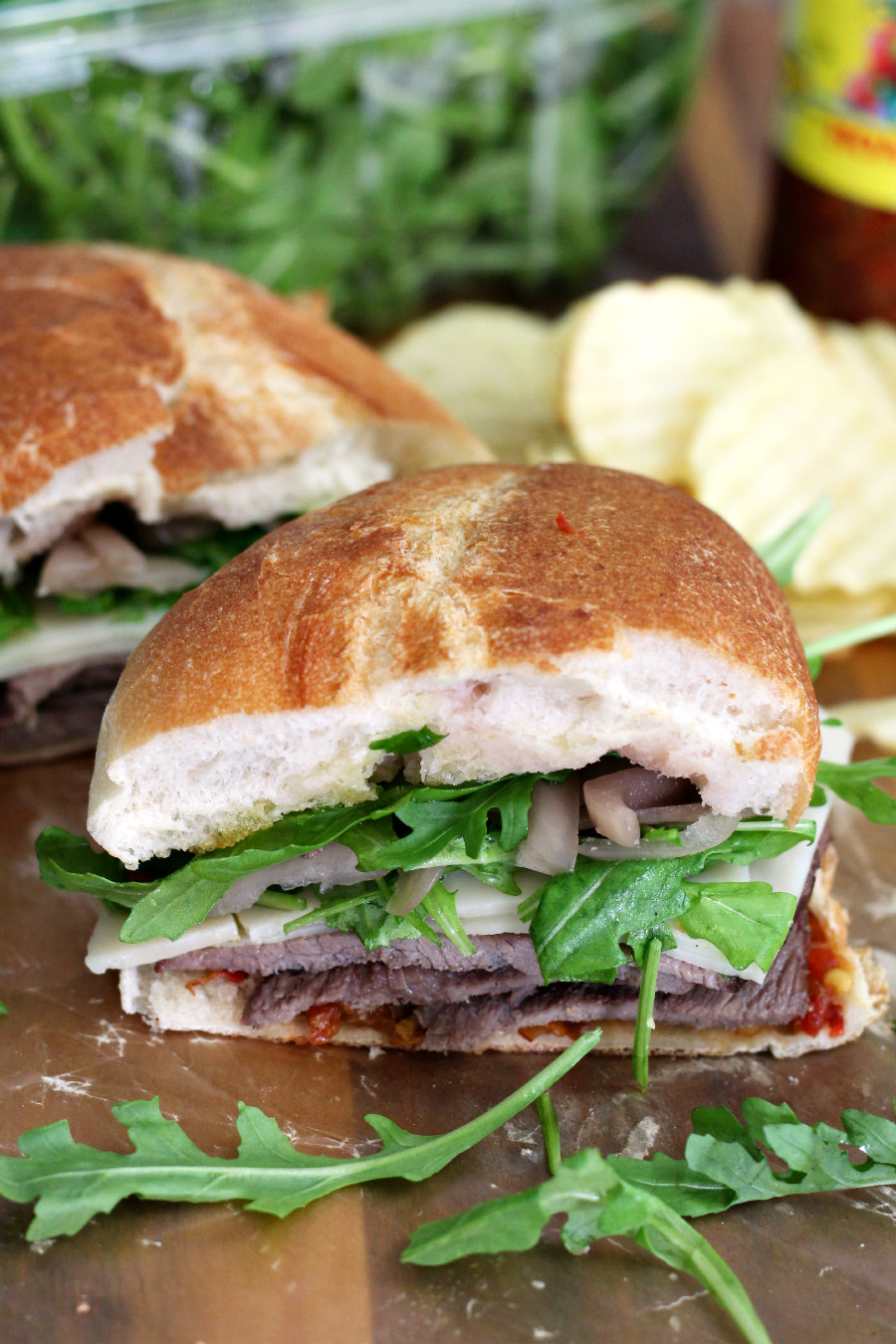 Roast Beef and Cherry Pepper Sandwich half sits on wooden board with other half of sandwich in partial view in background. Arugula, potato chips, and jar of pepper spread also in partial view.