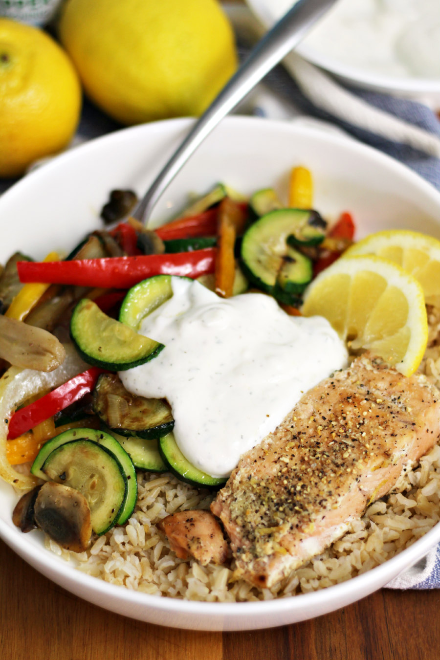 Lemon Pepper Salmon Rice Bowl sits on wooden board. Fork sits inside bowl. Lemons and bowl of yogurt dip in partial view in background.