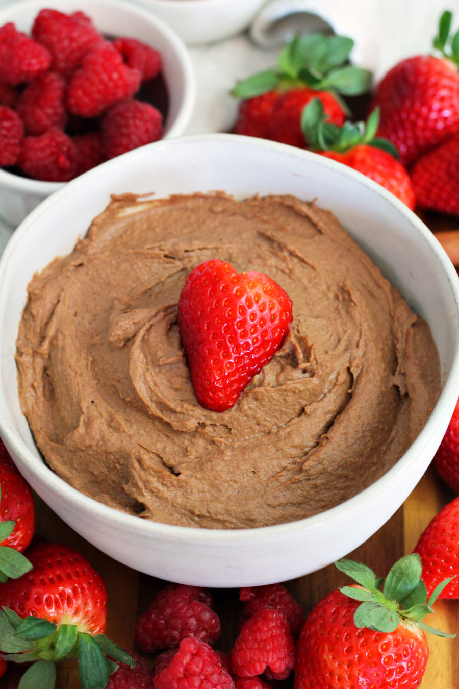 Chili Chocolate Hummus sits in serving bowl on wooden board. Heart shaped strawberry tops hummus. Strawberries and raspberries sit around bowl.
