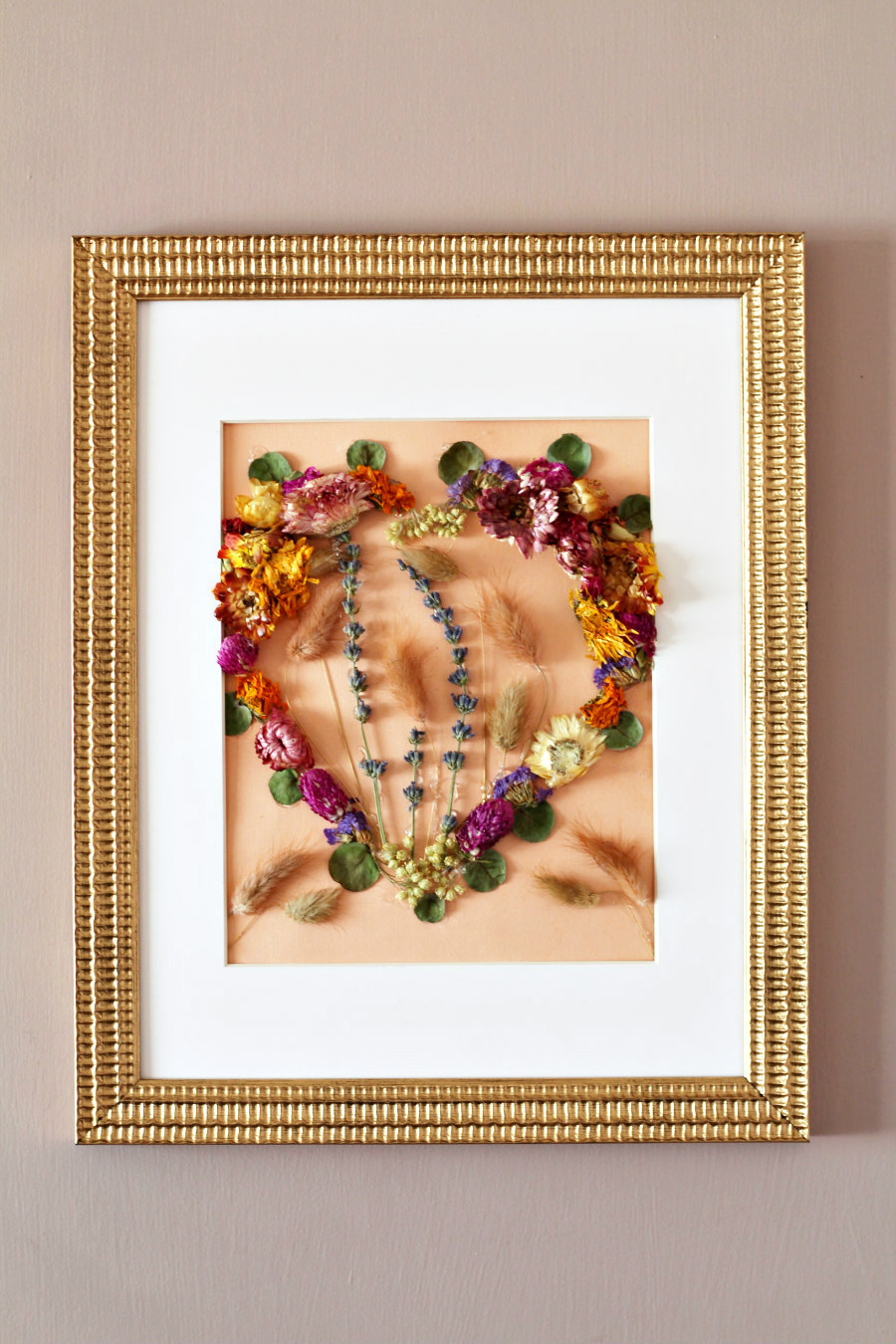 Dried flower Valentine's Day art in a gold frame hung up on the wall. Colorful dried flowers are arranged in a heart shape on peach colored cardstock.