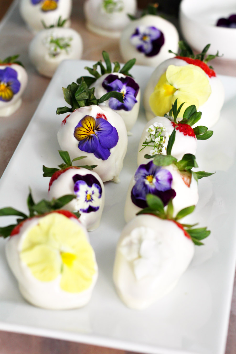 White Chocolate-Covered Strawberries with Edible Flowers sit on white serving plate. Other chocolate-covered strawberries and small white bowl in partial view in background.