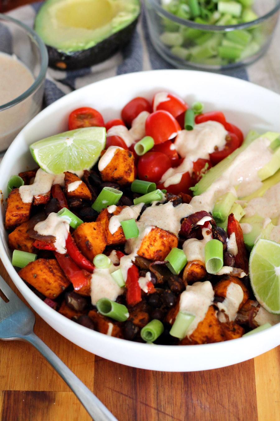 Cajun Sweet Potato and Black Bean Nourish Bowl sits in white serving dish on top of wooden board. Fork, bowl of yogurt sauce, halved avocado, and bowl of scallions also in partial view.