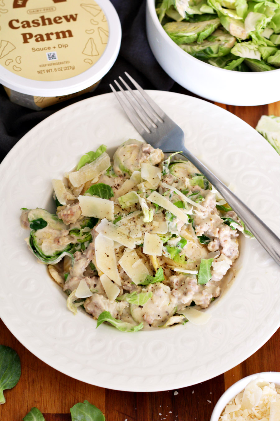 Butternut Squash Ravioli with Sausage and Shaved Brussels Sprouts in serving dish with fork on plate. Dish sits on wooden board. Cashew Parm container, bowl of shaved Brussels sprouts, and small bowl of shaved Parmesan also in partial view.