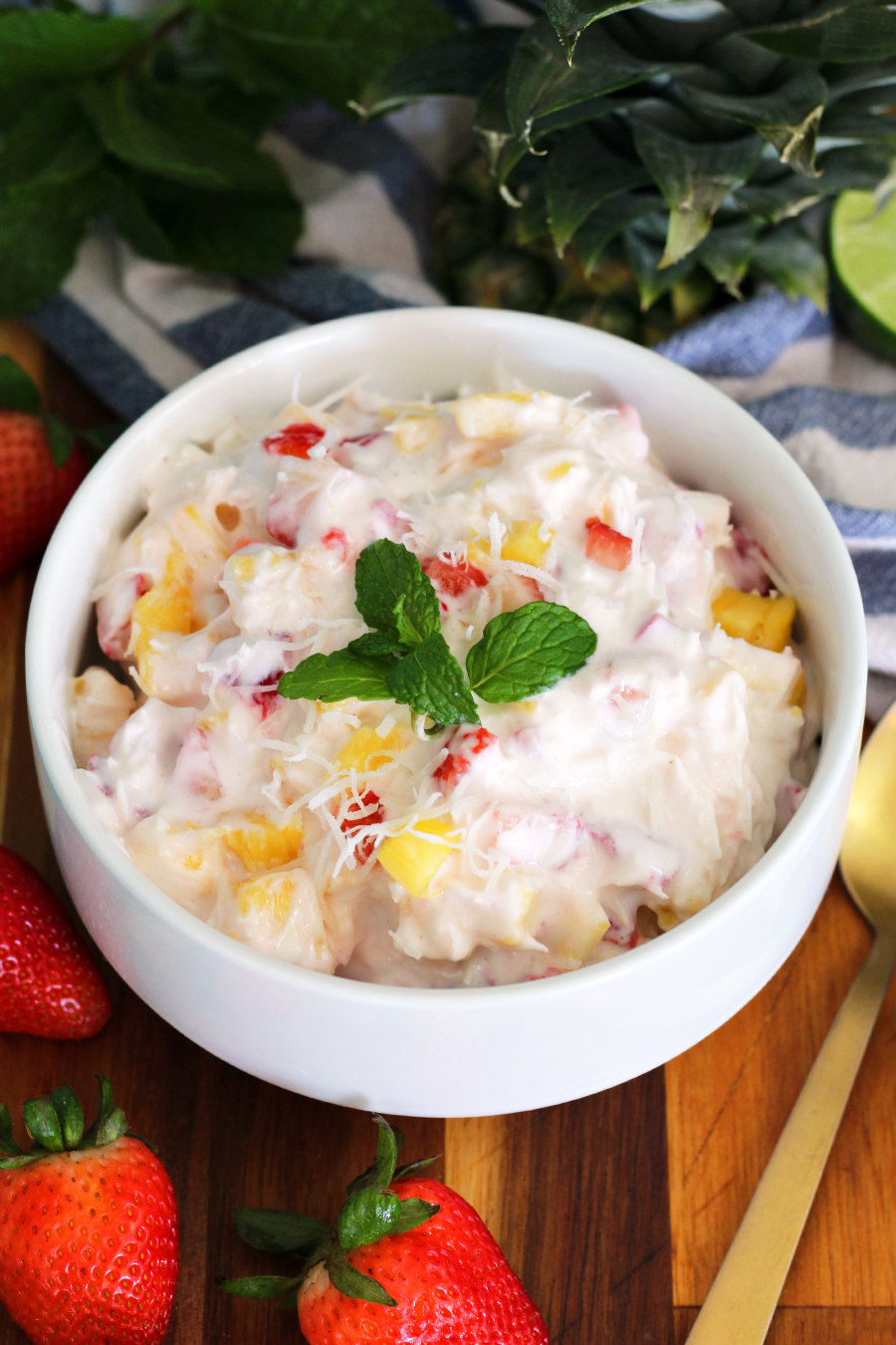 Tropical Strawberry Coconut Pineapple Yogurt Salad in white bowl sits on wooden board. Gold spoon, strawberries, mint leaves, pineapple top, and halved lime also in partial view.