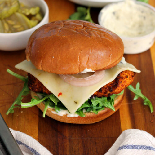 Hot honey chicken sandwich sits on wooden board with garlic sauce container, bowl of pickles, bowl of arugula, tongs, and white kitchen towel in partial view.