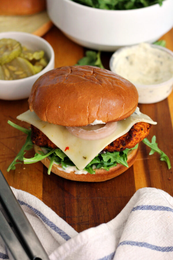 Hot honey chicken sandwich sits on wooden board with garlic sauce container, bowl of pickles, bowl of arugula, tongs, and white kitchen towel in partial view.