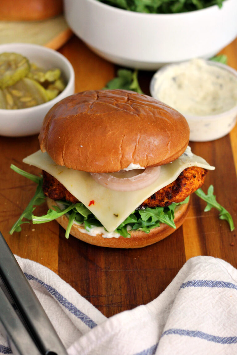 Hot honey chicken sandwich sits on wooden board with garlic sauce container, bowl of pickles, bowl of arugula, tongs, and white kitchen towel in partial view.