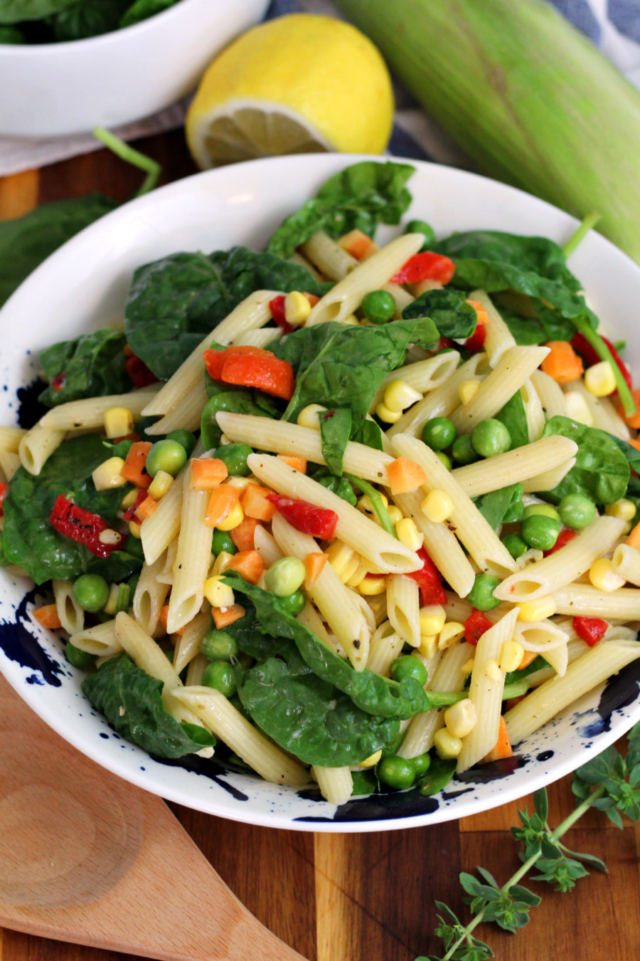 Vegetable Penne Pasta Salad in serving dish sits on wooden board. Wooden spoon, fresh oregano sprig, baby spinach, lemon, and ear of corn also in partial view.