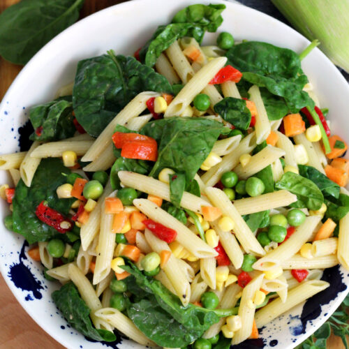 Top view of Vegetable Penne Pasta Salad in serving dish. Salad sits on wooden board. Wood spoon, baby spinach, lemon, ear of corn, and fresh oregano sprigs also in partial view.